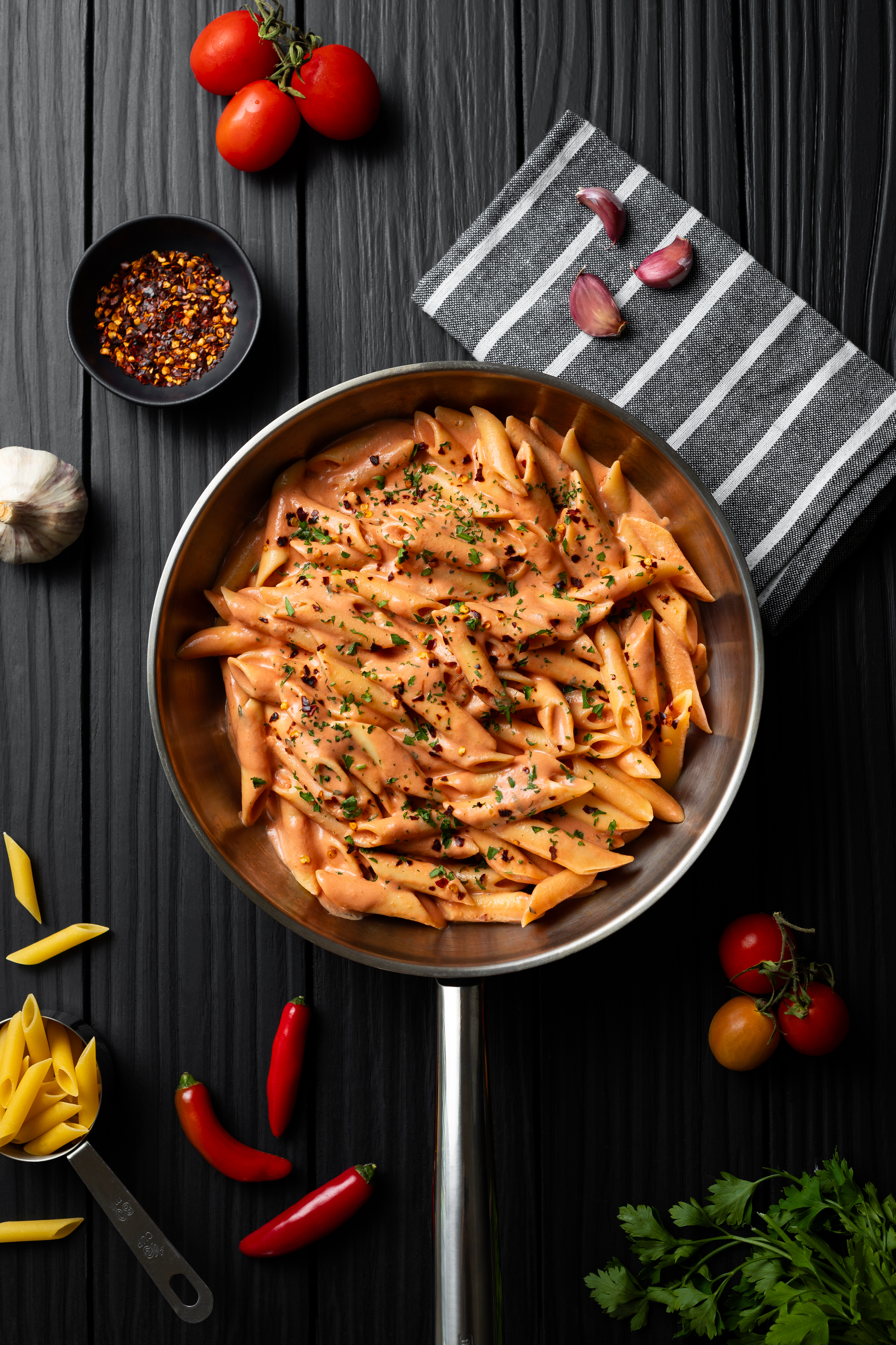 foto de um macarrão (penne alla vodka) numa frigideira servido com salsinha e pimenta calabresa. tomates, alho, salsinha e pimenta dedo de moça no cenário